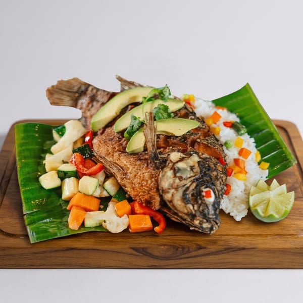 Whole fried fish with avocado, cilantro, rice, mixed vegetables, and lime on a banana leaf.